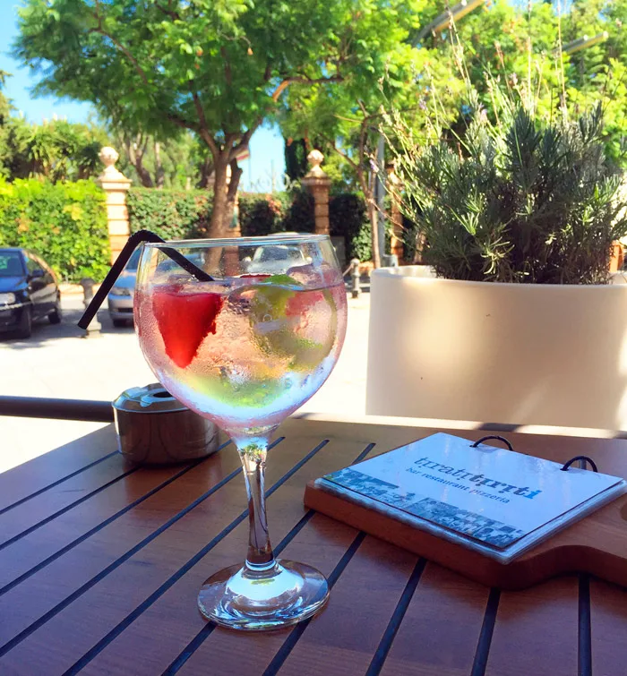 Copa de gin-tònic amb fruita a la terrassa, amb els arbres de la plaça al fons